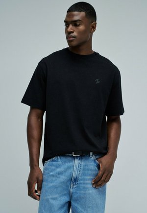 Homme portant un t-shirt noir uni et un jean bleu, regardant vers sa gauche avec une main dans la poche sur un fond gris uni.