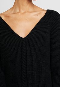 Žena ve černém žebrovaném pleteném svetru s výstřihem do V, který odhaluje její klíční kost a horní část hrudníku.