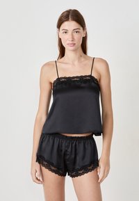Kobieta w czarnym satynowym topie na ramiączkach i szortach z koronkowym wykończeniem, stojąca na jednolitym jasnym tle.