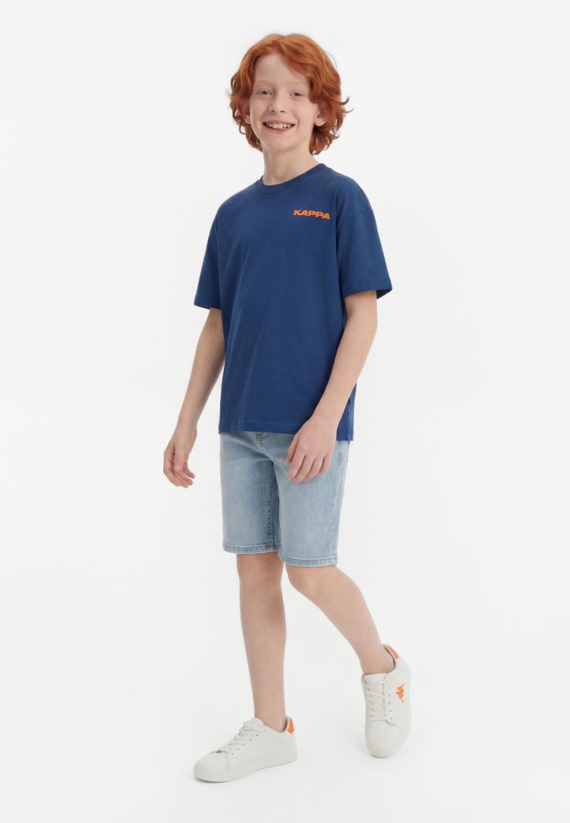 Garçon roux souriant debout, portant un t-shirt Kappa bleu marine, un short en jean clair et des baskets blanches avec des touches orange sur fond blanc.