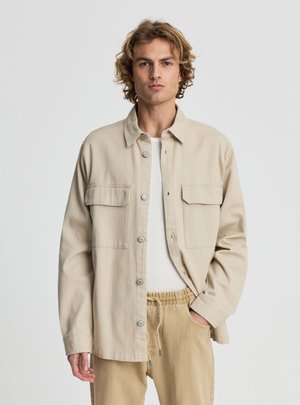 Młody mężczyzna o kręconych włosach, ubrany w beżową kurtkę na guziki na białą koszulę oraz beżowe spodnie z troczkami, stojący na jednolitym tle.