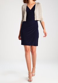 Navy blue dress with gathered front design, paired with a short, light gold cardigan. Model wears silver strappy sandals.