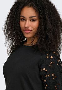 Woman with curly dark hair, wearing black top with floral lace cut-out sleeves, smiling softly against a light gray background.