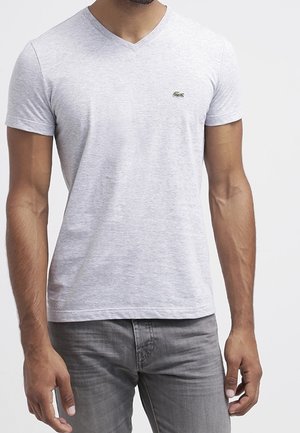 Homme portant un t-shirt gris clair à manches courtes et col en V avec un petit logo crocodile et un jean gris sur fond blanc.
