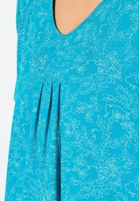 Turkos topp med V-ringning, blommig prägel i ljusare nyanser och vertikala veck för extra detaljer. Slät tygtextur.