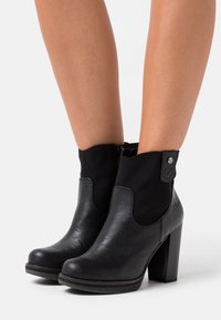 Botas de tobillo de cuero negro con tacones gruesos, llevadas sobre piernas desnudas, que cuentan con una cremallera lateral y un panel trasero texturizado.