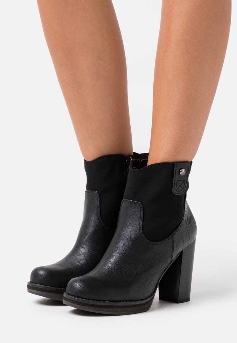 Botas de tobillo de cuero negro con tacones gruesos, llevadas sobre piernas desnudas, que cuentan con una cremallera lateral y un panel trasero texturizado.