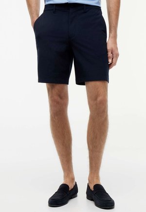 Man draagt marineblauwe op maat gemaakte shorts met zijzakken en zwarte loafers, staand tegen een effen witte achtergrond.