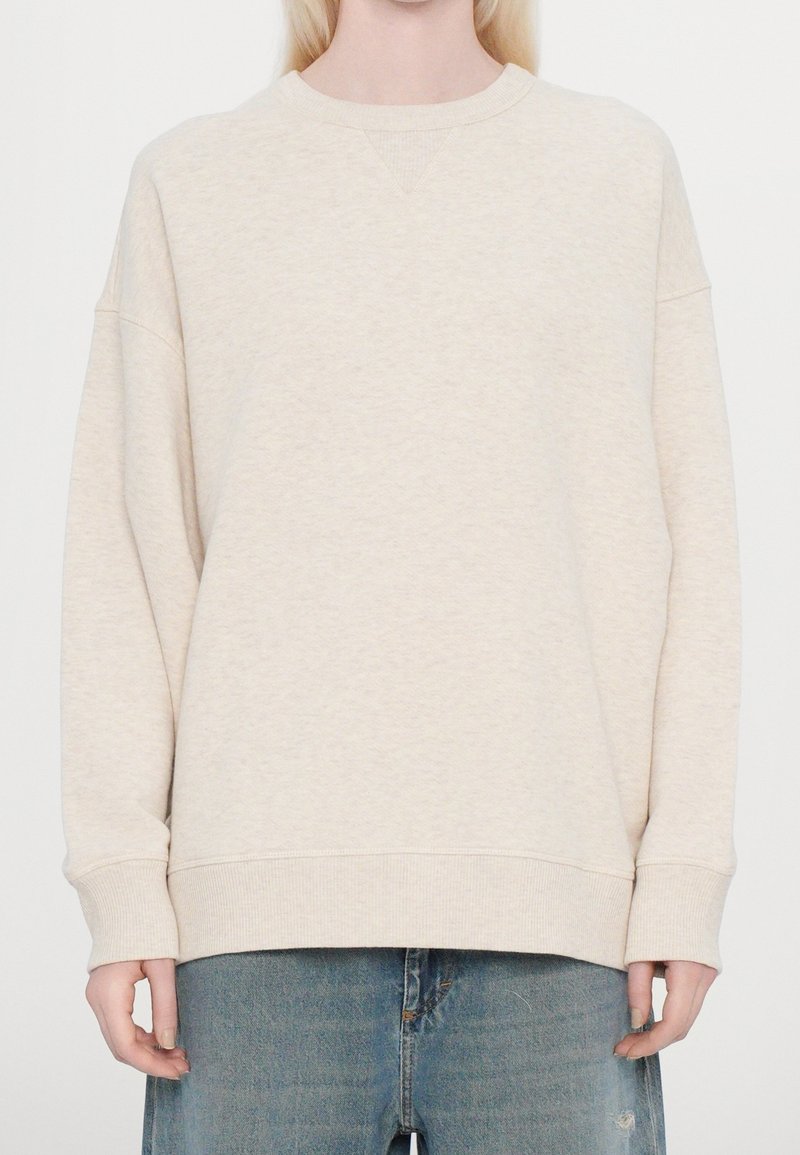 Personne portant un sweat-shirt ample beige clair à col rond et un jean en denim bleu, debout devant un fond blanc uni.