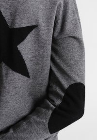 Grauer Strickpullover mit einem großen schwarzen Sternmuster und schwarzen Ellenbogenpatches. Die Textur wirkt weich und leicht strukturiert.