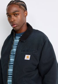 Junger Mann mit geflochtenem Haar, der eine schwarze Carhartt-Jacke über einem blau gestreiften Hemd vor einem schlichten weißen Hintergrund trägt.
