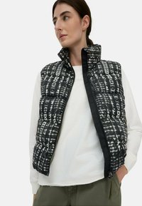 Zwart-wit gewatteerd vest met hoge kraag, voorzien van een geometrisch patroon, ritssluiting en zijzakken, gedragen over een wit shirt.