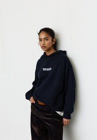 Granatowa bluza z kapturem z kieszenią kangurka, z białym logo "NEW STUDIO ROTTERDAM" na piersi. W połączeniu z ciemnymi sztruksowymi spodniami.