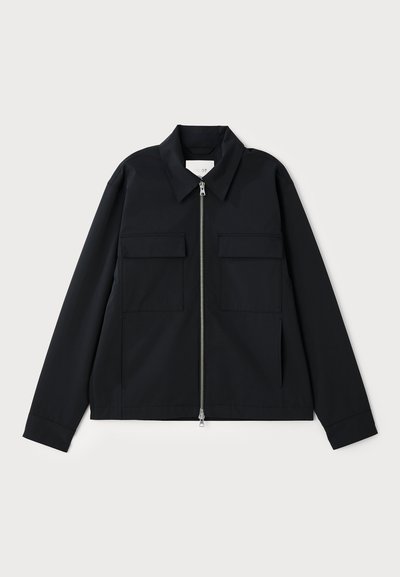 Chaqueta negra con cuello, cierre frontal de cremallera y dos bolsillos en el pecho. Confeccionada en un tejido suave y con un corte estructurado.