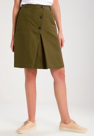 Kobieta ubrana w oliwkowozieloną spódnicę na guziki do kolan oraz beżowe sznurowane tenisówki z płótna, stojąca na tle białego tła.