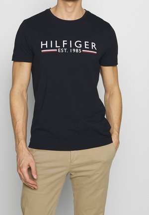 Mężczyzna w czarnej koszulce Tommy Hilfiger z napisem „HILFIGER EST. 1985”, w beżowych spodniach, stoi na jednolitym tle.