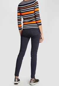 Maglione a righe con strisce arancioni e blu navy, abbinato a jeans skinny scuri e sneakers metallizzate, visto da dietro.