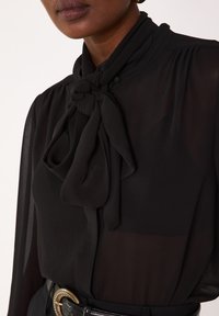 Blouse noire avec un col lavallière, tissu fluide, épaules plissées et texture transparente, associée à une ceinture noire avec boucle dorée.