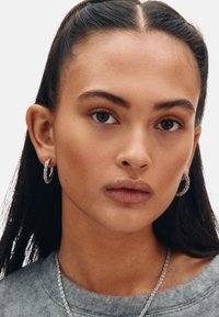 Hõbedast kõrvarõngad, tekstuurse disainiga, mida kannab modell, kellel on sirged tumedad juuksed ja hall särk, valge taustaga.