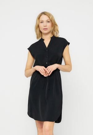 Blondide juustega naine, kes kannab lühikeste varrukatega musta nööpidega ees kleiti, seisab käed õrnalt vööl kokkupandud valge tausta ees.