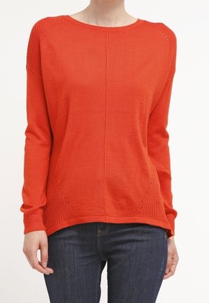 Femme portant un pull en maille à manches longues de couleur orange vif avec des détails discrets de coutures, associé à un jean en denim bleu foncé.