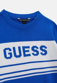 Blauer Strickpullover mit einem weißen Streifen und "GUESS" in fetten blauen Buchstaben, runder gerippter Halslinie und glatter Textur.