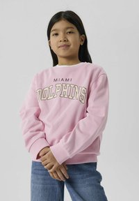 Rozā kapuci krekls ar lieliem burtiem "MIAMI DOLPHINS", riboti aproces un apakšmalu, kopā ar zila džinsu biksēm. Mīksta auduma tekstūra.