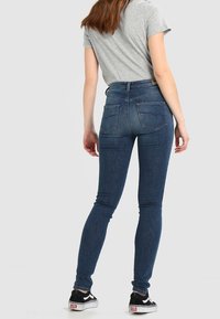 Jeans de tiro alto en azul, con un ajuste estrecho, hechos de mezclilla, con bolsillos traseros y una textura suave, combinados con zapatillas negras.