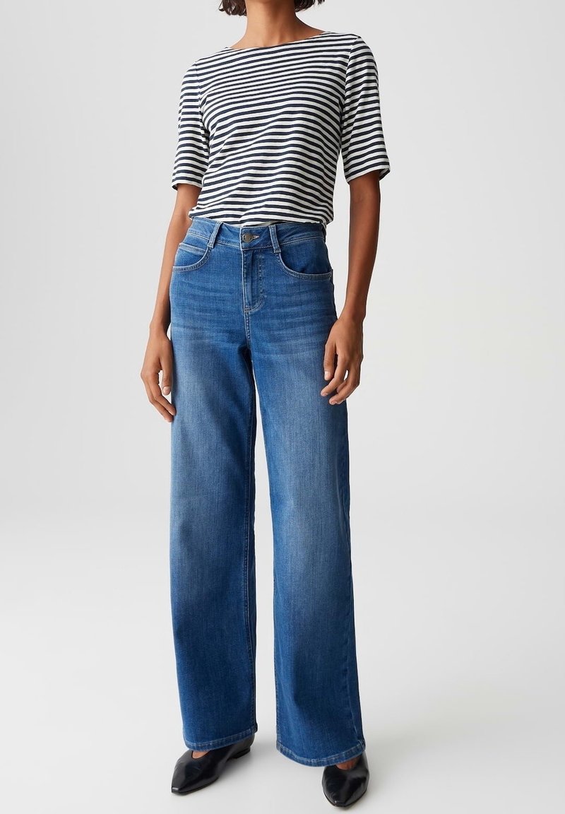 Femme portant un haut à manches courtes rayé noir et blanc, un jean bleu taille haute à jambes larges et des chaussures noires pointues, debout devant un fond uni.