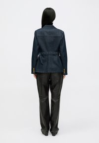 Giacca di jeans scura con vita a cintura, dettagli in bottone dorato e vestibilità su misura. Indossata sopra pantaloni in pelle nera, visibile da dietro.