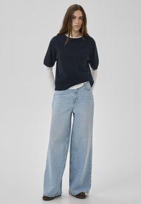 Marineblaues Kurzarm-Strickoberteil, übereinander getragen auf einem weißen Langarmshirt, kombiniert mit hellblauen Jeans mit weitem Bein. Minimale Details und weiche Textur.