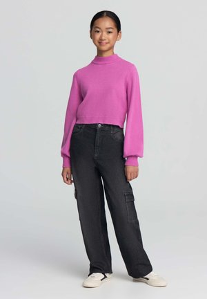 Giovane che indossa un maglione rosa brillante, pantaloni cargo neri larghi e sneakers bianche, in piedi su uno sfondo chiaro e semplice.