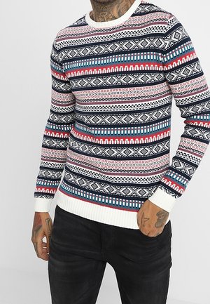 Mężczyzna ubrany w kolorowy sweter w wielokolorowy wzór z białymi mankietami oraz czarne spodnie, pokazujący tatuaże na rękach i szyi.