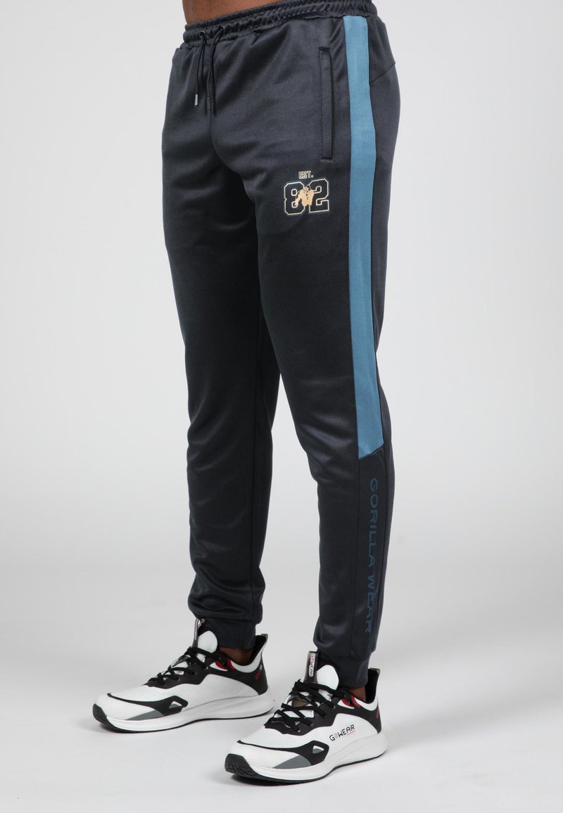 Zwarte sportbroek met een blauwe zijkant en gouden logo. De stof is glad. Heeft een elastische tailleband en taps toelopende benen.