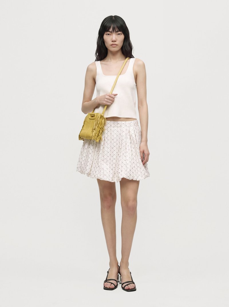 Une femme porte un haut blanc sans manches, une jupe à motifs, des sandales noires et un sac bandoulière jaune à franges, sur un fond uni.