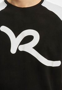 Juodas marškinėliai su dideliu baltu stilizuotu "VR" logotipu. Rankovės baltos, su lygiu tekstūru, kontrastuojančiu su tamsiu audiniu.