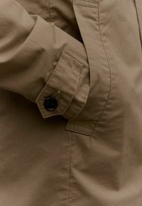 Giacca marrone con una superficie liscia, dotata di polsini con bottoni, tasca laterale e dettagli trapuntati. Design semplice e funzionale.