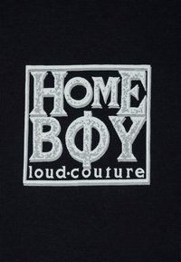 Schwarzer Stoff mit einer weißen Grafik, die den Text "HOME BOY" und "loud.couture" in einem rechteckigen Design zeigt. Der Text hat einen abgenutzten Effekt.