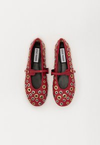 Ballerines en cuir rouge avec des découpes circulaires et des œillets métalliques. Dotées d'une doublure intérieure noire et d'une sangle à boucle assortie.
