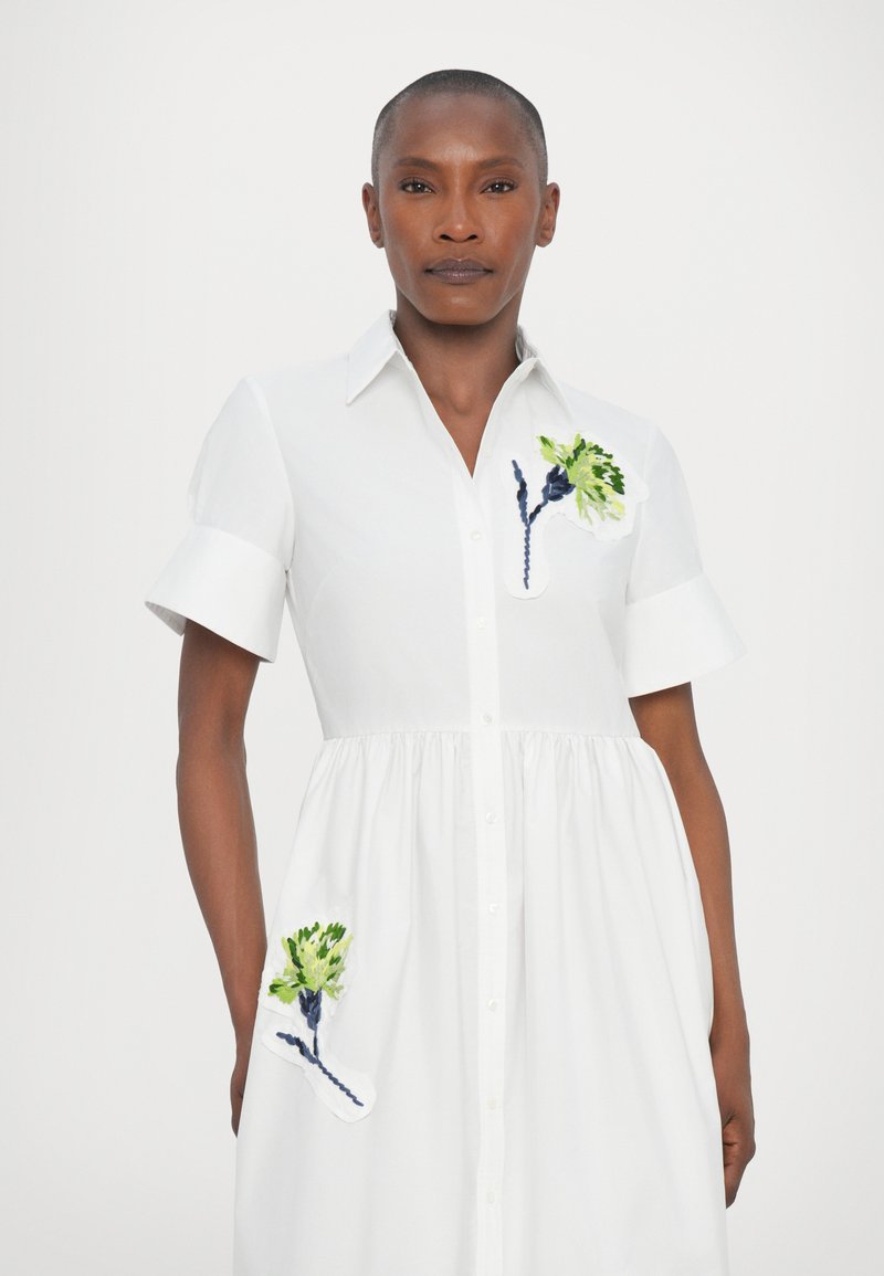 Abito in cotone bianco con maniche corte, chiusura a bottoni e gonna arricciata; presenta ricami floreali verdi e blu navy sul petto e sulle tasche.