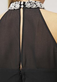 Vestido negro semitransparente de halter en la parte posterior con un collar de perlas, cerrado con dos grandes botones de perla sobre piel desnuda.