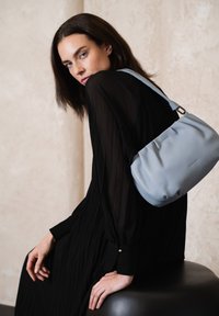 Helleblaue Lederschultertasche mit glatter Textur, lässiger Form und einem einzigen Tragegurt, getragen über einem fließenden schwarzen Plisseekleid.