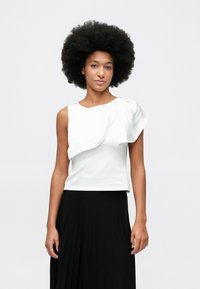 Young woman with natural afro hairstyle wearing a sleeveless white top with ruffled detail and a black pleated skirt, standing against white background.