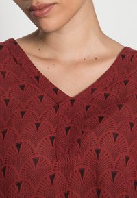 Nahaufnahme des Halses und des unteren Gesichts, das ein rotes, gemustertes V-Ausschnitt-Oberteil mit schwarzen, fächerförmigen Motiven trägt.