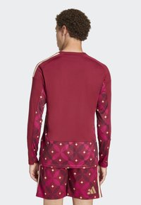 Majica dugih rukava za sport u boji burgundac s geometrijskim ružičastim uzorkom. Značajke uključuju kontrastne pruge na ramenima i prilagođeni dizajn.