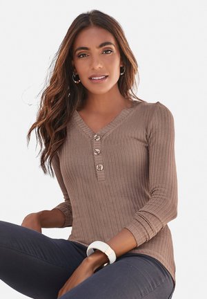 Top marron à manches longues côtelé avec un décolleté en V et trois boutons à l'avant. Porté avec un pantalon sombre et un bracelet de couleur claire.