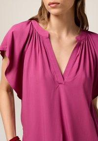 Blusa fucsia con scollatura a V, spalle plissettate e maniche corte. Realizzata in tessuto leggero e morbido, presenta una vestibilità comoda e una texture liscia.