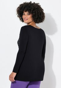 Schwarzes Langarmshirt mit Sternmuster auf der Vorderseite, aus weichem Stoff und mit rundem Halsausschnitt. Das Model trägt es mit lila Hosen und zeigt einen lässigen Schnitt.