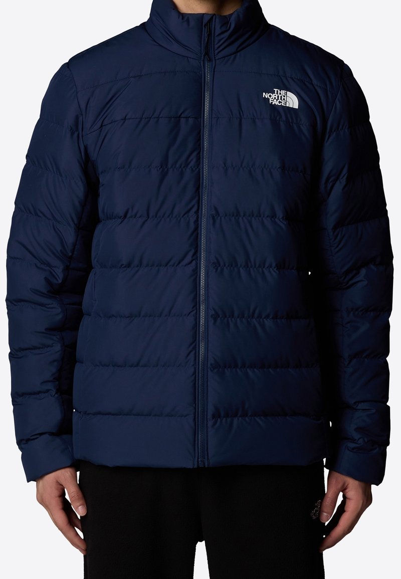Veste matelassée isolante bleu marine avec col montant, motif matelassé horizontal, fermeture éclair intégrale et logo sur la poitrine gauche.