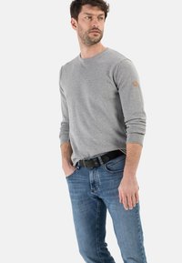 T-shirt à manches longues gris avec col rond et poignets côtelés, associé à un jean en denim bleu et une ceinture noire. Design simple avec un patch logo sur la manche.